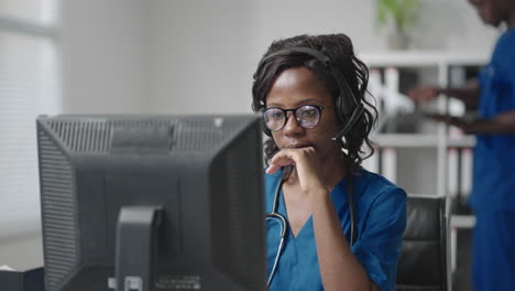 A-black-woman-doctor-wearing-headphones-sits-at-a-table-with-a-computer-and-takes-calls-from-patients-looks-at-their-medical-records-and-enters-them-into-the-clinic-schedule